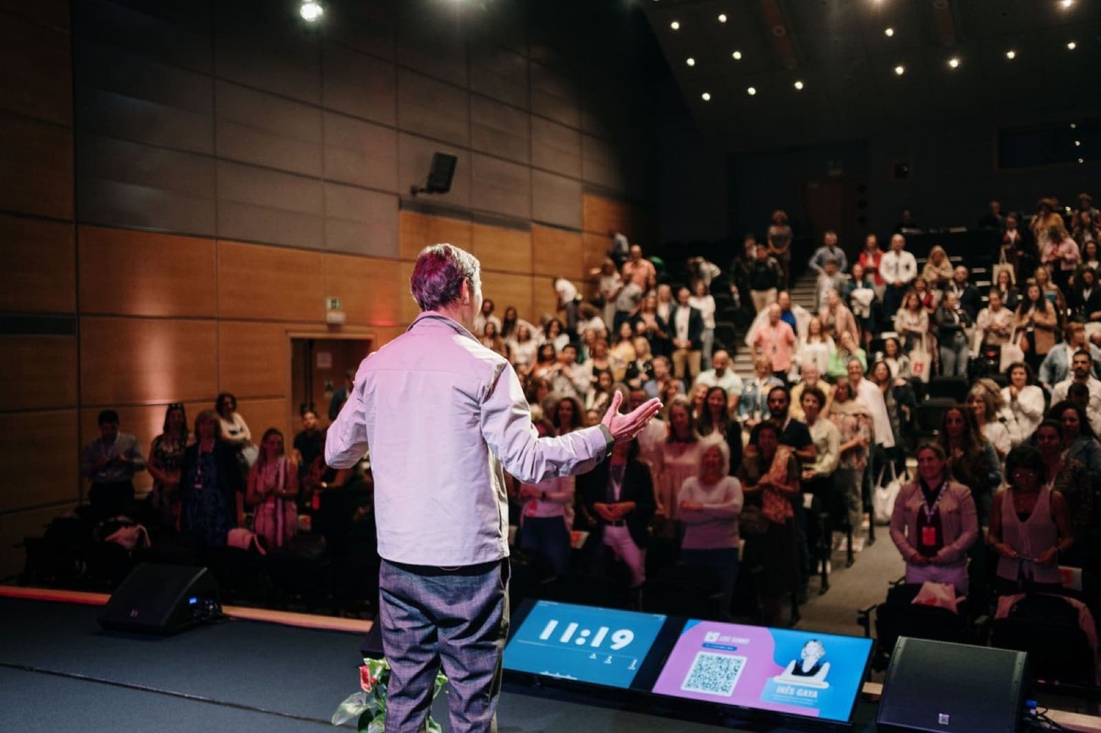 A speaker seen from behind opens his arms to a standing auditorium audience at Love Summit