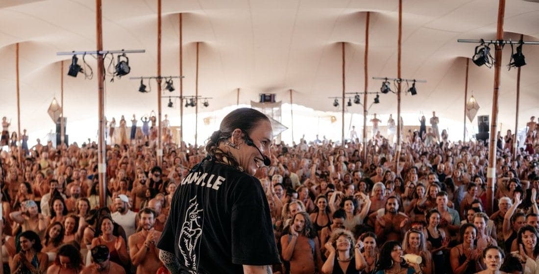 A facilitator on stage faces a full tent at Medicine Festival, guests with hands raised